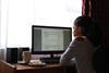 An image showing a girl studying a computer screen, sat at a desk, with a mug and biscuits to her left, all in front of a window
