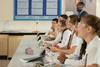 High school students wearing safety glasses and school uniform watching a demonstration in a science classroom. They are smiling and engaged.