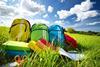 Brightly colour backpacks lined up on grass in a sunny field with books and scissors beside them 