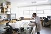 A picture showing a female technician wearing a white lab coat in a school laboratory with a textbook, microscope and fume cupboard visible