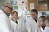 A teacher and younger high school students in a practical science class. The students are in uniform and they are all wearing lab coats and safety goggles. One girl is raising her hand.