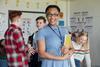 A female teacher holds a textbook and smiles; behind her a group of students in casual clothes talk between themselves
