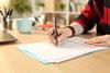 A student's hands holding a pen and writing on an exam paper on a desk; a notebook, mug and laptop are visible in the background