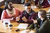Three students sitting at a table with a tablet, pens, paper, having a discussion