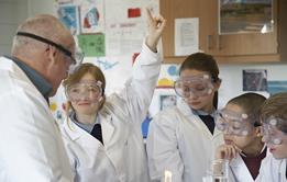 A teacher and younger high school students in a practical science class. The students are in uniform and they are all wearing lab coats and safety goggles. One girl is raising her hand.