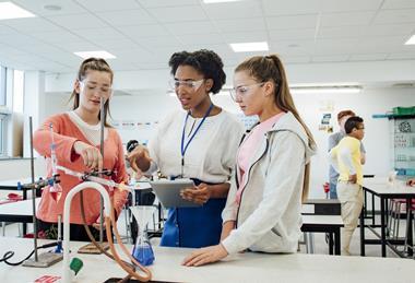 A photograph of two students and their teacher discussing a chemistry experiment, with a variety of apparatus set up in front of them