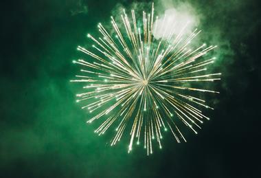 A photograph showing a green firework exploding in the night sky, surrounding by smoke