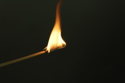 A close-up photograph of a lighted wooden splint against a black background