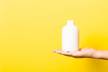 A photograph showing an open hand holding a plain white shampoo bottle in front of a yellow background