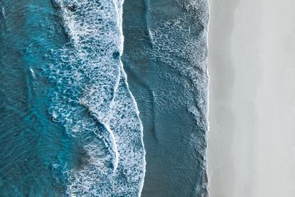 An overhead drone photograph of a neutral grey sandy beach and waves from the sea