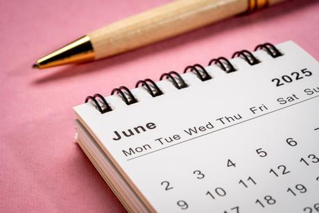 Photograph of a small spiral desktop calendar showing June 2026 on a pink background with a pen in view