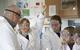 A teacher and younger high school students in a practical science class. The students are in uniform and they are all wearing lab coats and safety goggles. One girl is raising her hand.