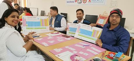 Three teachers sit around a desk holding up posters of the periodic table of elements