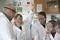 A teacher and younger high school students in a practical science class. The students are in uniform and they are all wearing lab coats and safety goggles. One girl is raising her hand.