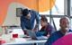 A male teacher bends down to discuss some work with a student, who is sitting at a desk with a notebook and laptop open in front of her