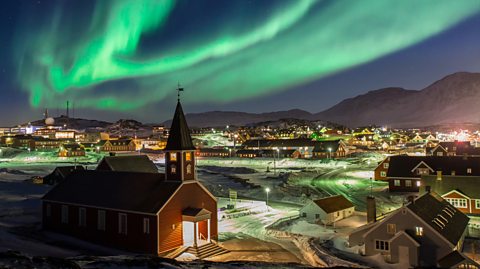 Northern Lights above Nuuk, Greenland. At the forefront of the photo is a brown church, surrounded by houses and lights in the city in the distance. Above, the Northern Lights provide a bright green haze to the night sky