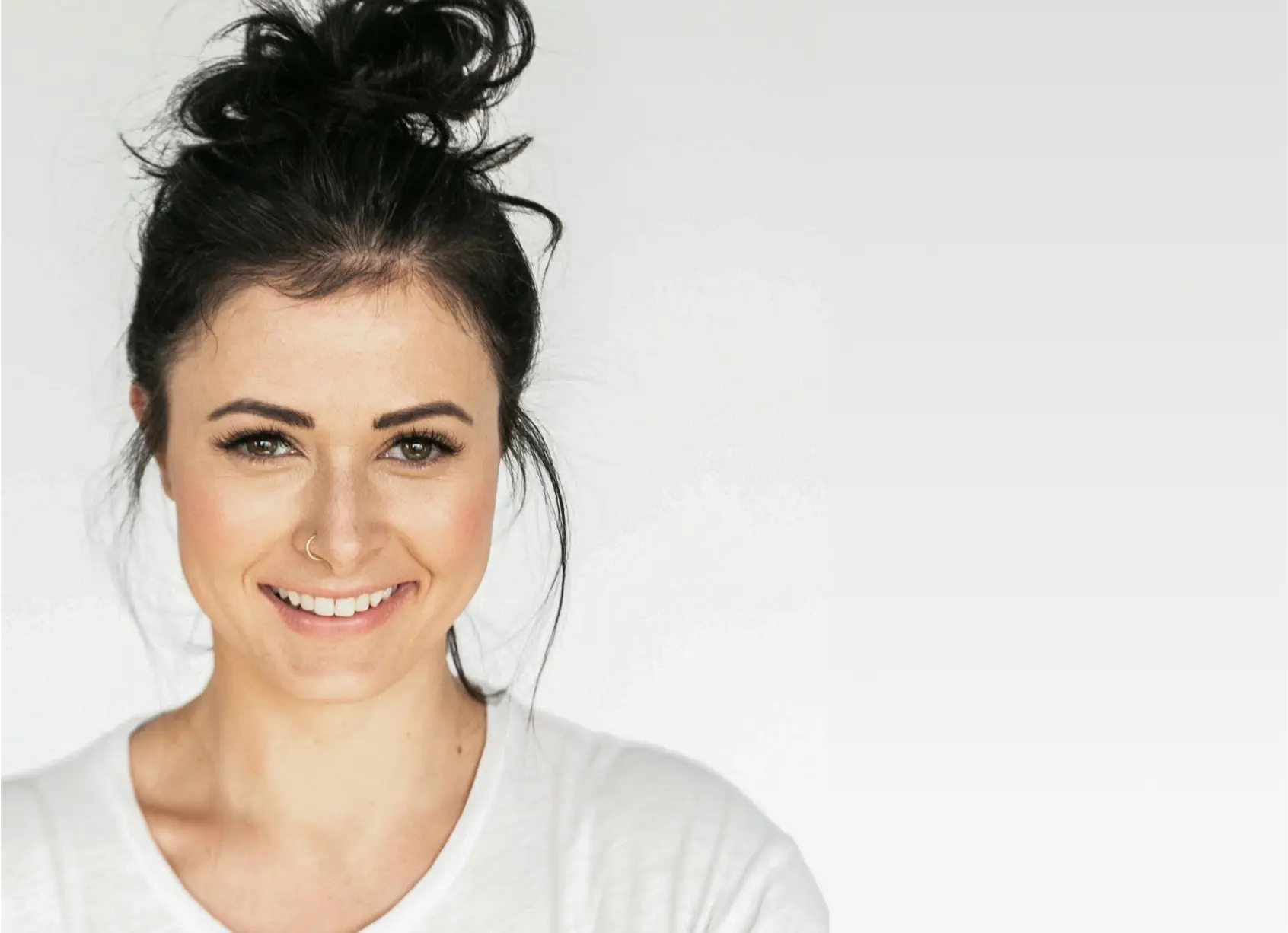 A young woman with dark hair in a messy bun, wearing a white shirt and a nose ring, smiles at the camera against a plain, light gray background.
