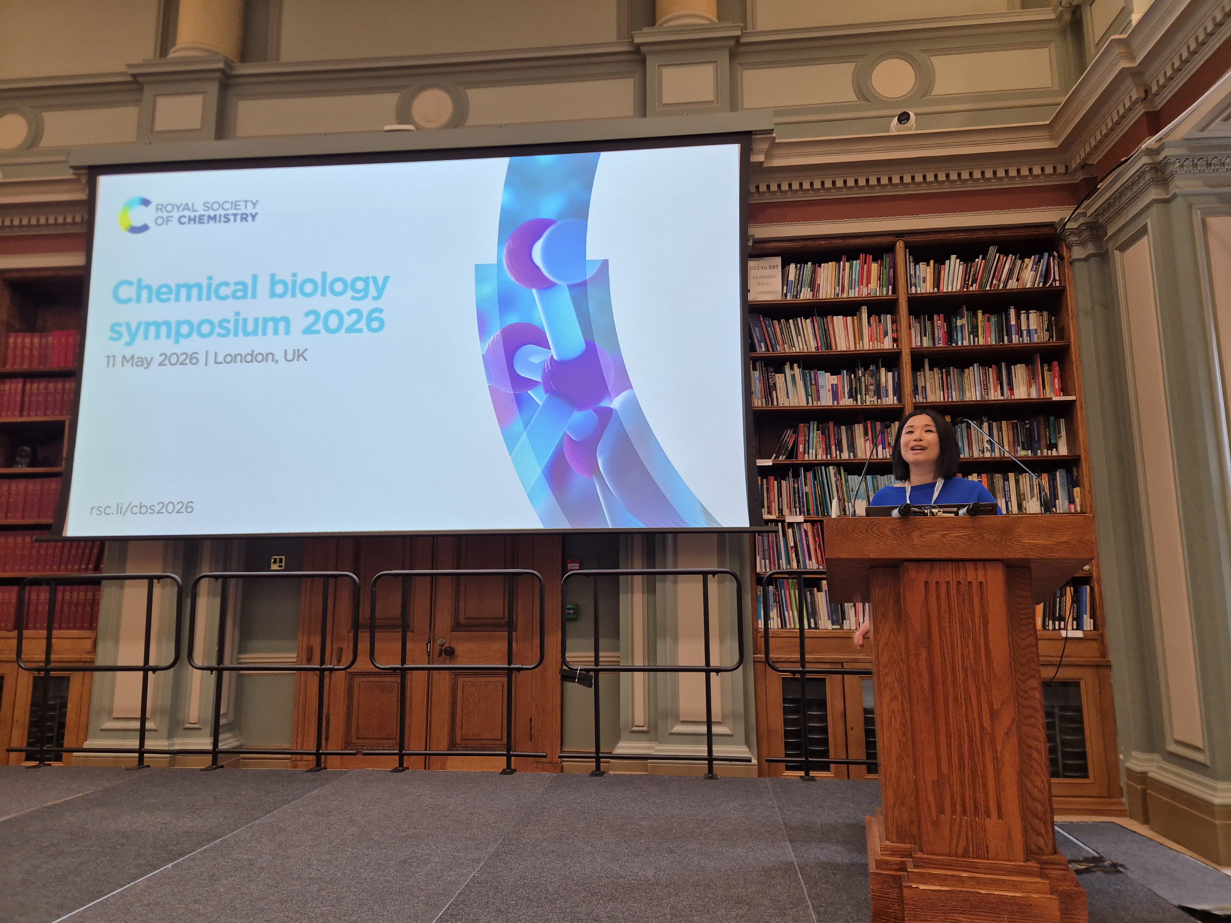 Professor Akane Kawamura stands behind a lectern to the right of the image, with a screen reading Chemical Biology Symposium 2026 on the other side of the imagerge screen