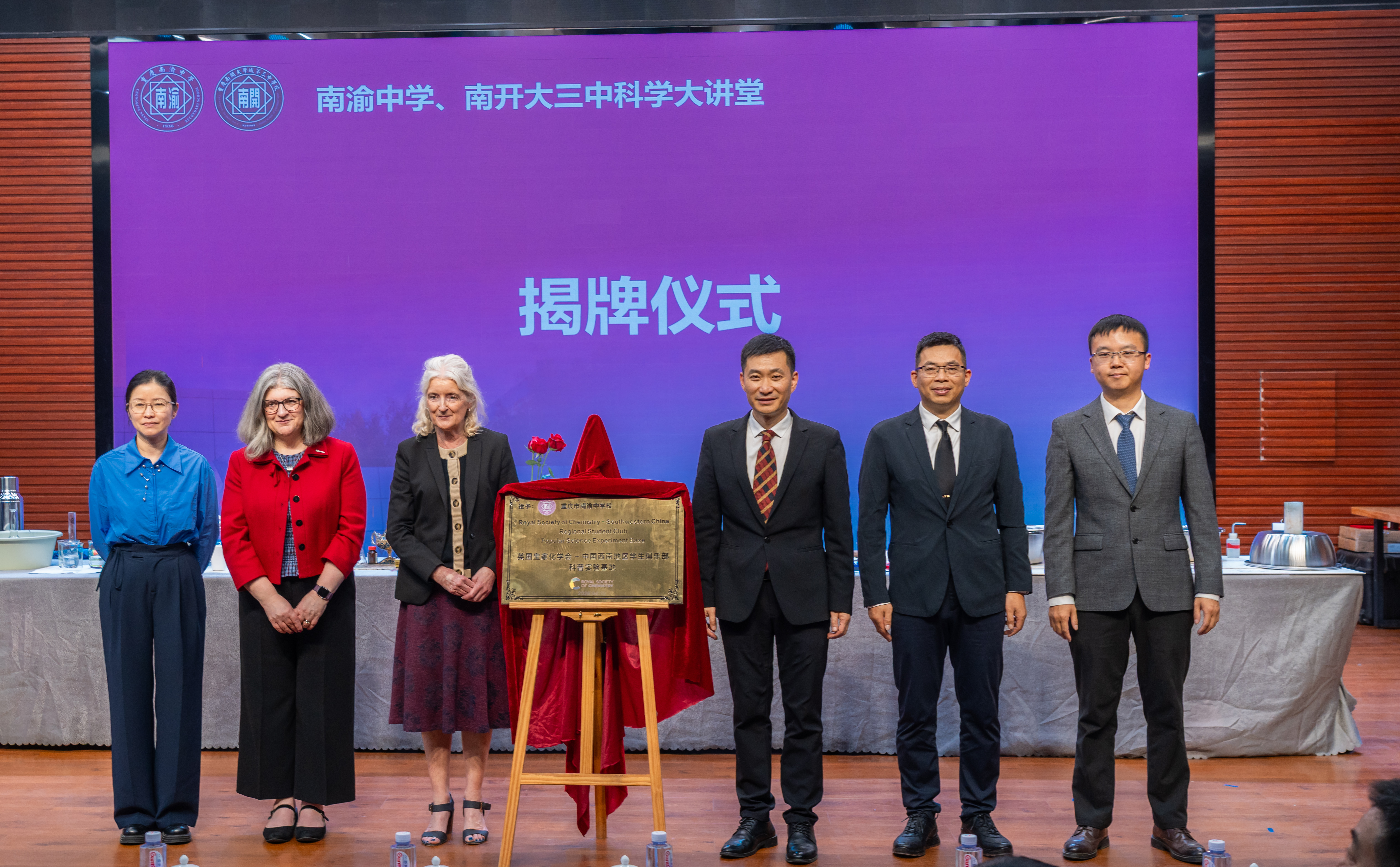 Three Royal Society of Chemistry representatives, including Dr Helen Pain and Dr Annette Doherty (second and third left), stand to the left of a golden plaque on an easel, with three men to the right