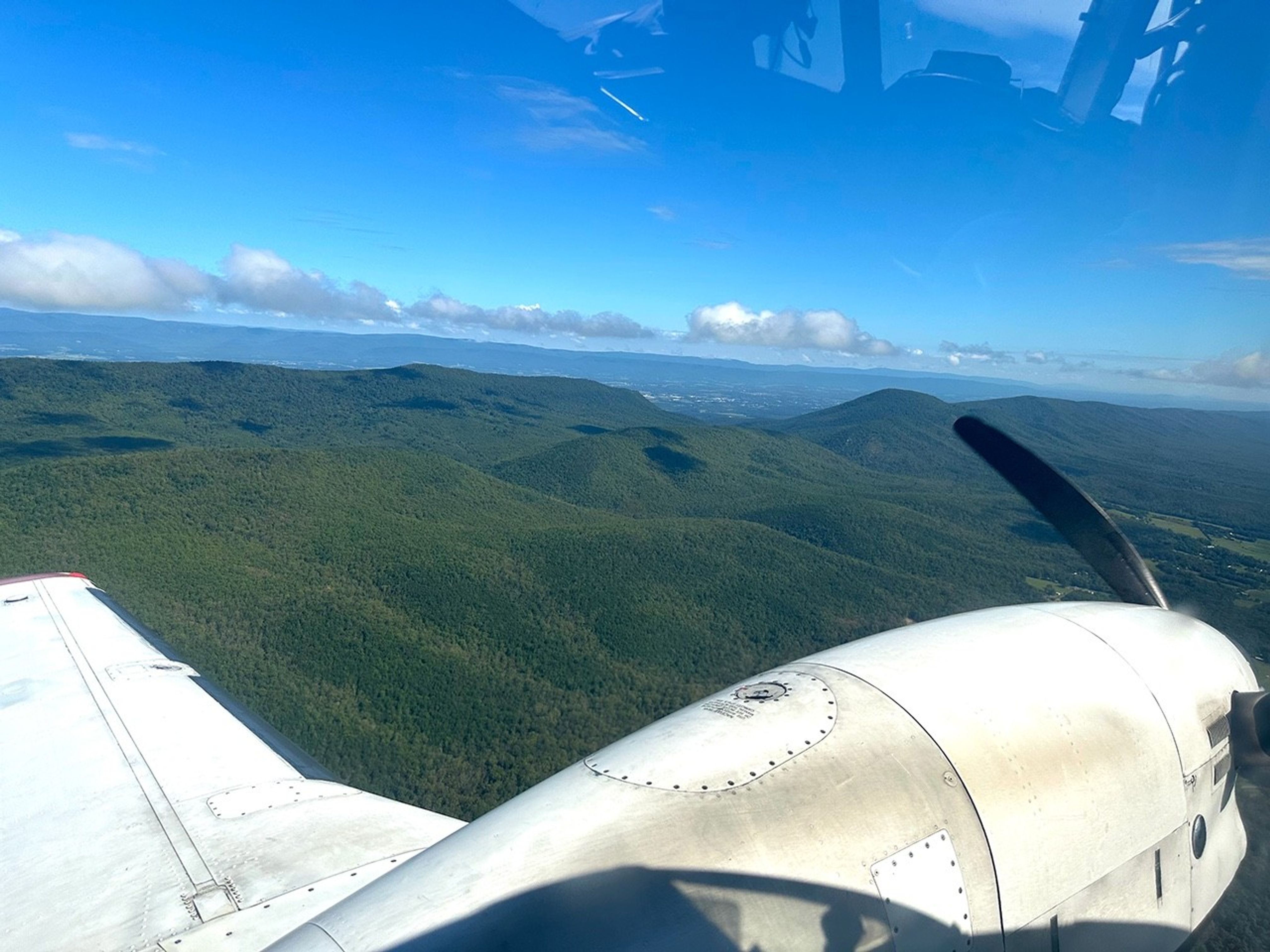 An aerial view of Shenandoah, showing green mountains and land. The horizon separates the bright blue sky from the land. At the top of the image is a reflection of inside the aircraft, showing this image was taken through a window. At the bottom of the image is a white wing of a plane and the engine and propellor of the same plane.