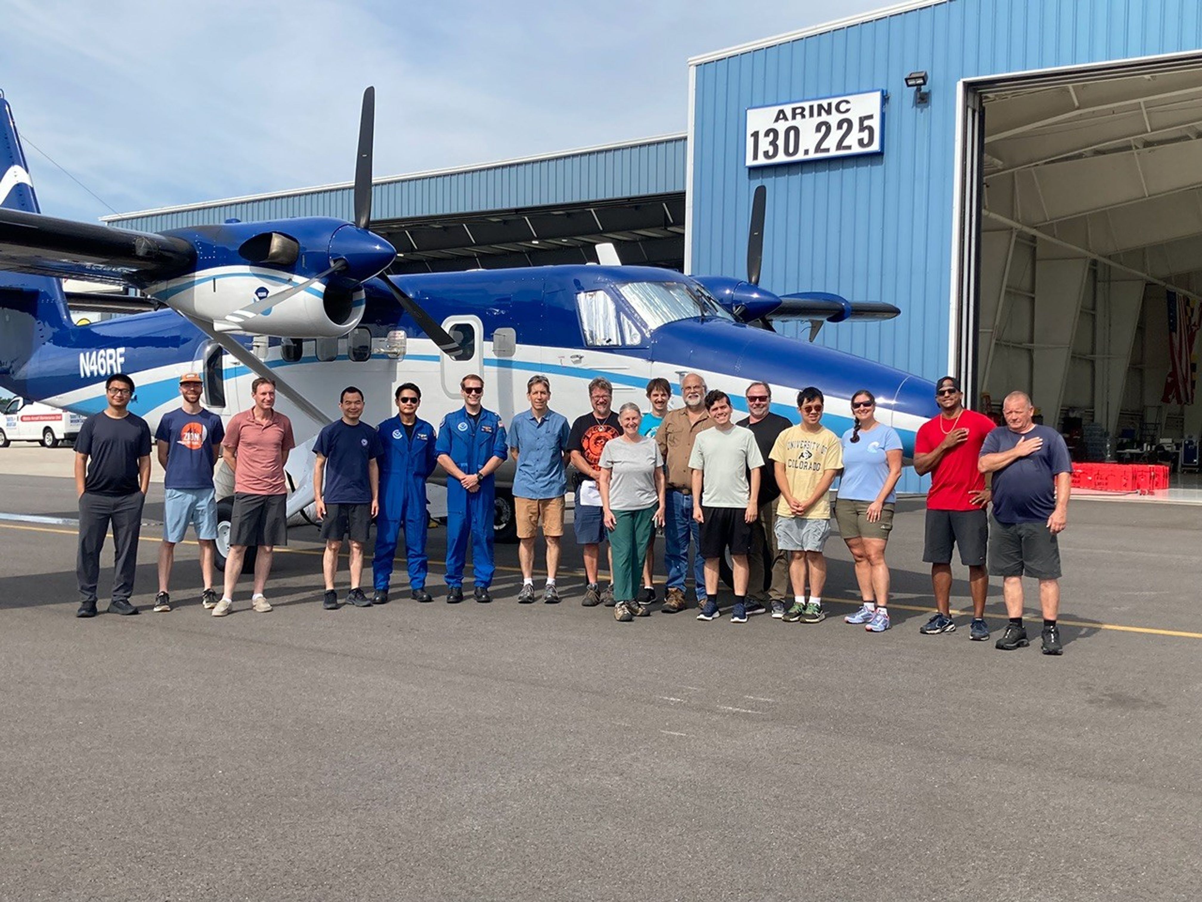 A group of seventeen people stand in a line in front of a blue aircraft with propellors. They all smile at the camera, which is taking a picture of them from a distance.