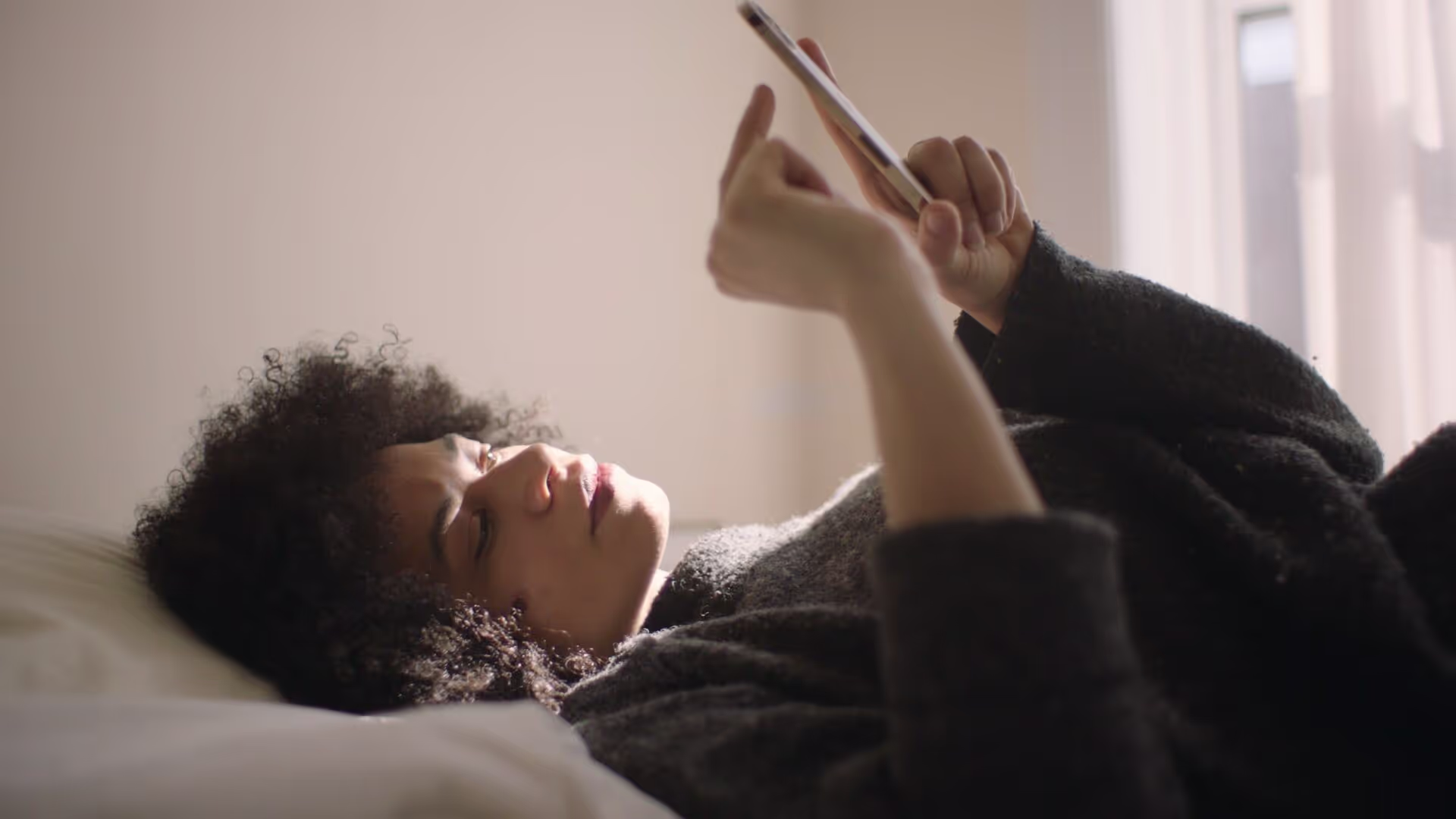 Person lying on a bed, holding and looking at a smartphone with both hands. Light from a window illuminates the scene.