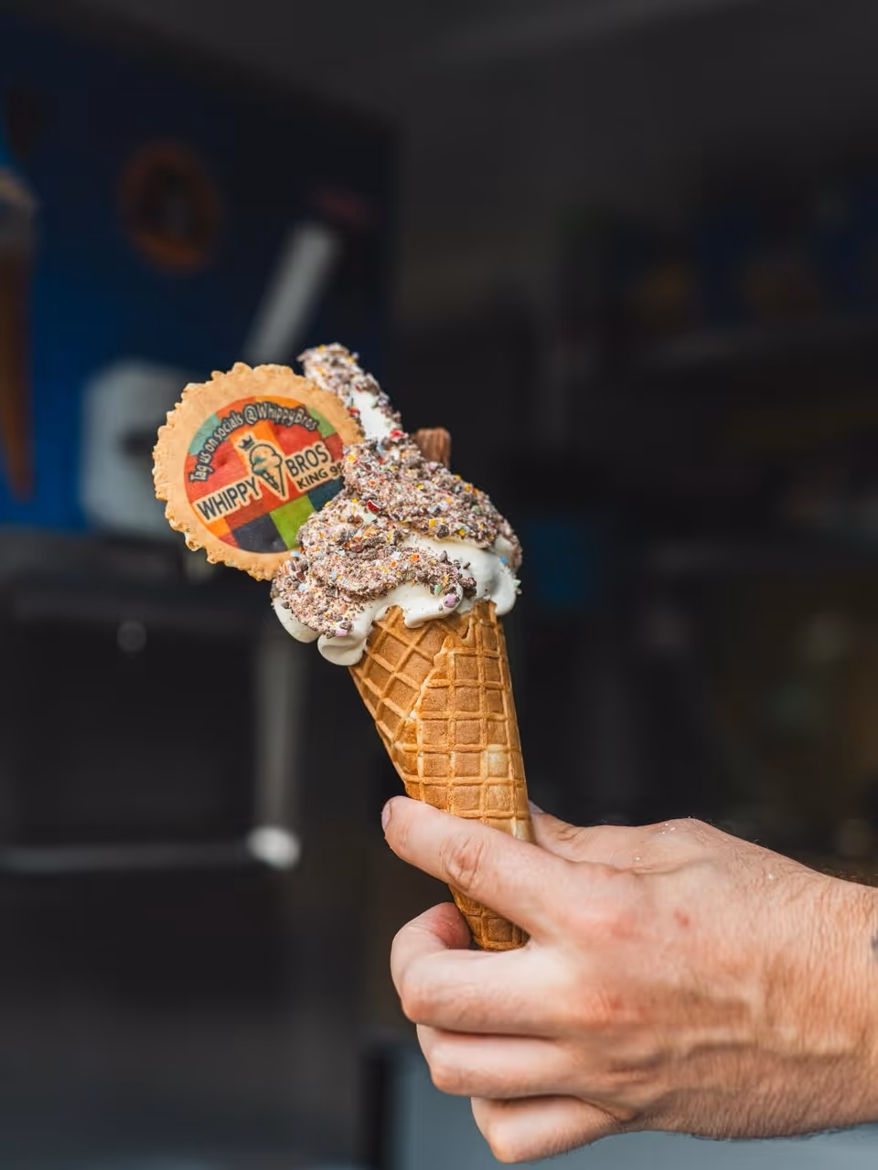 Hand holding an ice cream cone topped with sprinkles and a wafer cookie branded Whippy Bros.