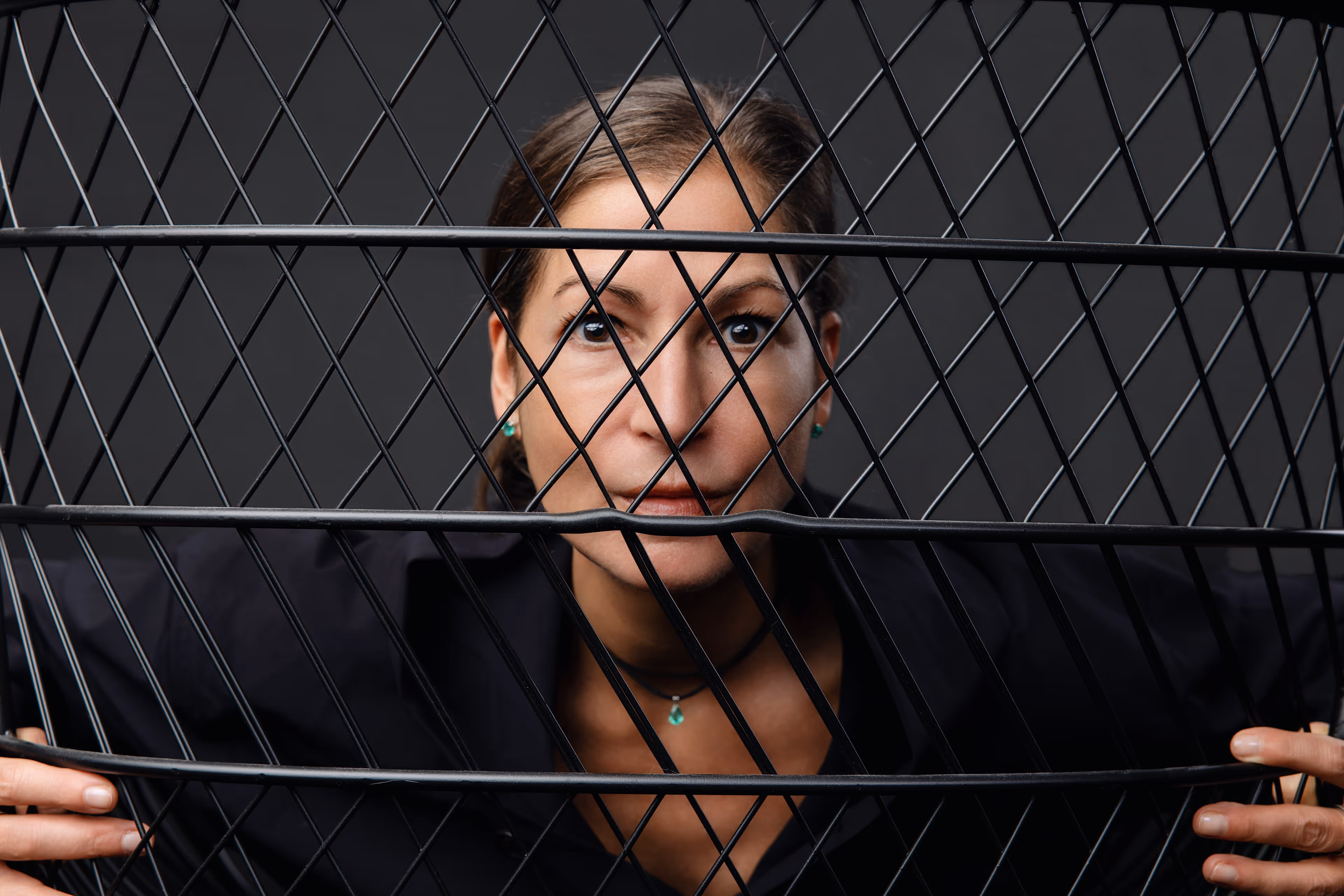 Frau blickt durch ein schwarzes Drahtgitter, hält es mit beiden Händen fest.