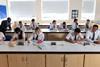 High school students wearing school uniform and safety glasses do written work in a school lab. The chemistry teacher is helping some of the students.