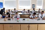 High school students wearing school uniform and safety glasses do written work in a school lab. The chemistry teacher is helping some of the students.