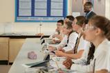 High school students wearing safety glasses and school uniform watching a demonstration in a science classroom. They are smiling and engaged.