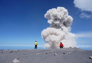 Two people stand near a large ash cloud rising from ash laden ground and against a sunny blue sky