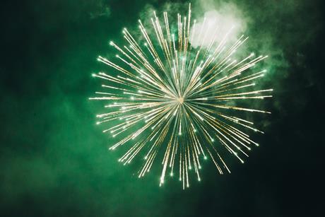 A photograph showing a green firework exploding in the night sky, surrounding by smoke