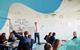 A male teacher stands at the front of a class pointing at equations on a whiteboard, while uniformed students listen and one raises their hand
