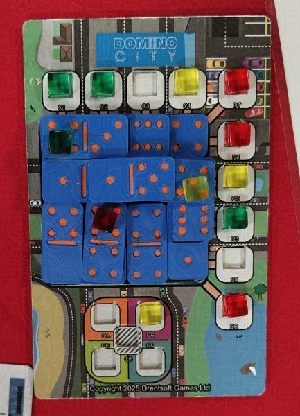 Domino City board on a Playtest UK red table at Paradice Worthing. A 3 player game between red, green and yellow players. Blue dominoes with orange pips are stacked on the board.