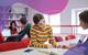 Two students sit opposite each other at a desk in a library, discussing a book open between them and pointing at a paragraph with their pens