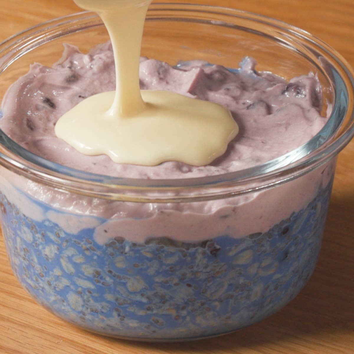White chocolate being poured over a bowl of blueberry-flavored oats in a glass jar