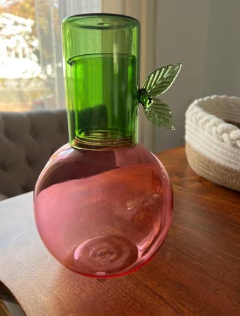 A decorative glass vase with a round base and tall neck, featuring a leaf detail, placed on a wooden table beside a woven basket
