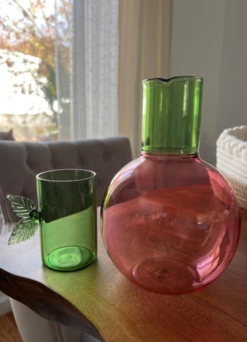 Green glass cup with leaf handle next to a round pink glass pitcher with a green spout, placed on a wooden table