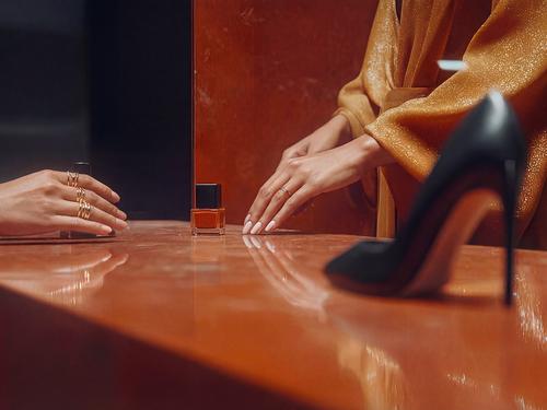 Les mains de deux femmes qui s’échangent un flacon de vernis à ongles orange sur un comptoir orange brillant, avec au premier plan une chaussure à talon haut noire.