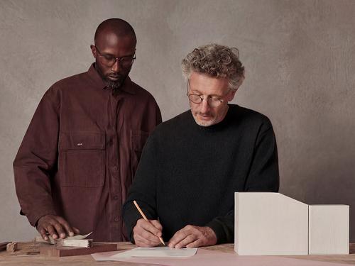Two architects, one older and one younger, reviewing plans and writing notes in an office setting with architectural models.