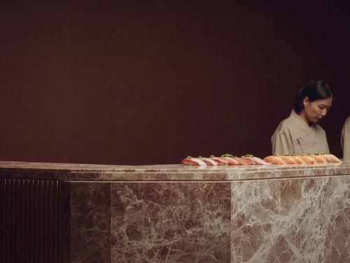 A sushi chef standing behind a marble counter displaying rows of expertly prepared sushi (sashimi and nigiri).