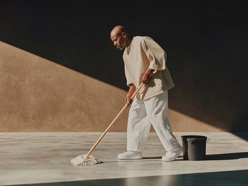 A bald man in light-colored clothing mops a large indoor floor area next to a dark bucket, with strong light and shadow contrast.