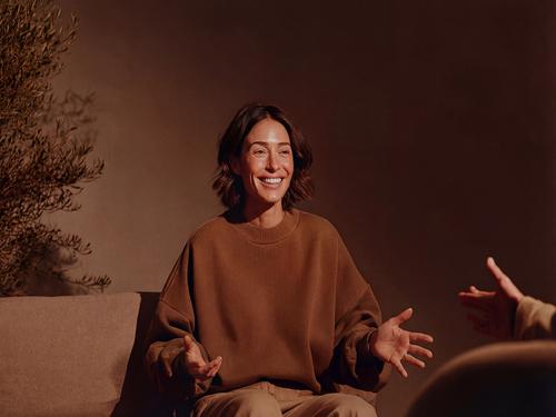 A smiling female coach or therapist in a brown sweater sitting on a couch, gesturing with open hands to an unseen client during a session.