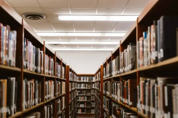 Rows of tall bookshelves filled with books line a library aisle, leading toward a distant wall with more shelves. Overhead fluorescent lights illuminate the space, where Book Ban Resources are available for those seeking information and support.