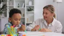 A woman therapist works with a young African American boy while he does some drawing.