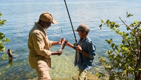 Two fishermen holding fishing poles alongside greenery and a blue lake.