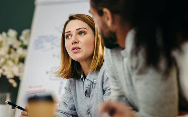 Two individuals engaged in a discussion in front of a blurred whiteboard with diagrams, suggesting a collaborative or educational setting