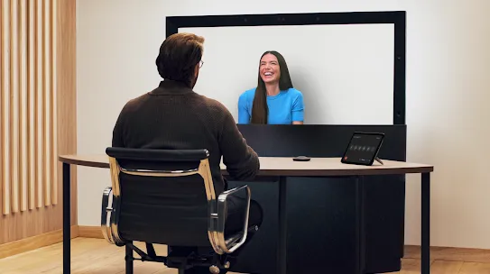 Two people use Google Beam. The person on the screen smiles at the person in the foreground.