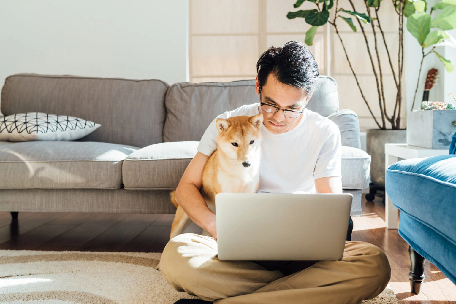 A man sits cross-legged on the floor in a bright living room, using his laptop to read a Hexclad case study. His Shiba Inu sits close beside him, both looking at the screen. A gray sofa, plants, and sunlight are in the background.